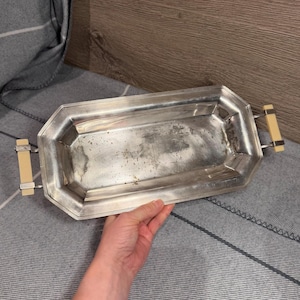 May include: A rectangular silver-toned metal serving tray with beveled edges and two light-coloured rectangular handles attached to the sides. The tray features a polished metallic surface with visible signs of wear and tarnishing throughout the base.