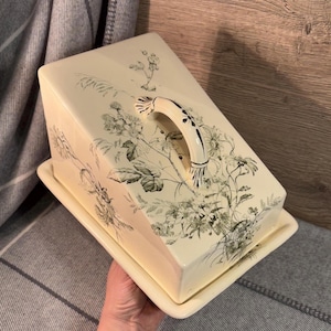 Peut inclure: Un dôme à fromage en céramique vintage avec un motif floral dans les tons de vert et de blanc. Le dôme a une poignée et une base séparée.