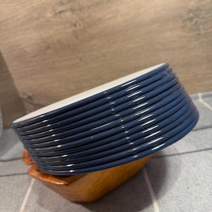 Pode incluir: Uma pilha de dez pratos redondos com bordas azul-escuras e centros brancos. Os pratos est&atilde;o inclinados e apoiados num pequeno suporte de madeira castanho-claro. As bordas dos pratos s&atilde;o lisas e brilhantes, com um design circular uniforme.