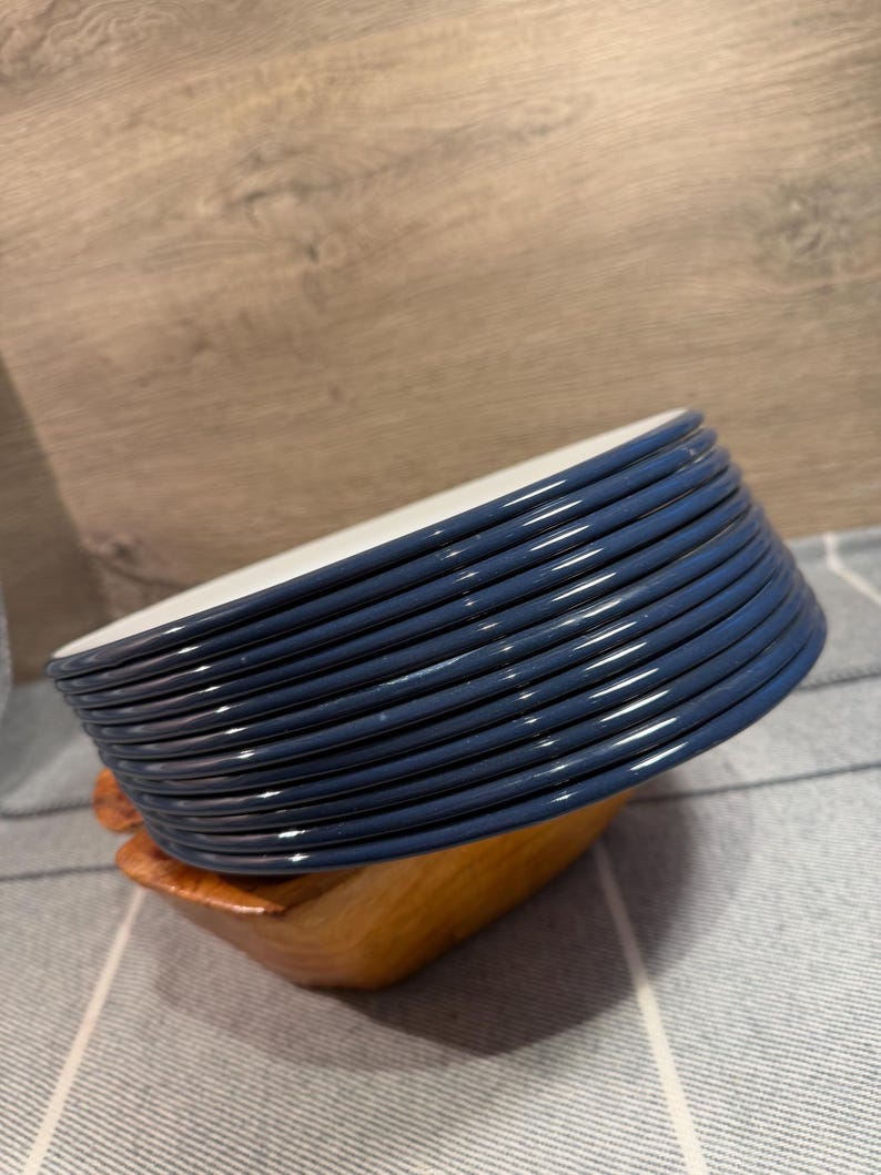 Pode incluir: Uma pilha de dez pratos redondos com bordas azul-escuras e centros brancos. Os pratos est&atilde;o inclinados e apoiados num pequeno suporte de madeira castanho-claro. As bordas dos pratos s&atilde;o lisas e brilhantes, com um design circular uniforme.