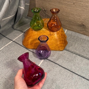 May include: Four small, colorful glass vases. Three vases sit on a wooden base: green, orange, and purple. A fourth, magenta vase is held in a hand. The vases have a teardrop shape and are transparent.