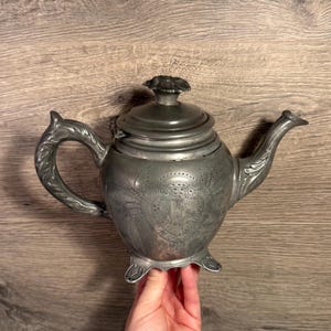 May include: A pewter teapot with detailed floral and scrollwork. The teapot has a curved handle, a bird-shaped spout, and a decorative lid. It stands on three small feet. The teapot is a classic design.