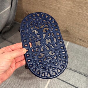 Puede incluir: Un salvamanteles de hierro fundido azul marino con forma ovalada y un intrincado diseño calado. El salvamanteles presenta un patrón floral y de volutas, y está diseñado para proteger las superficies de la cocina caliente.