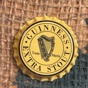 Puede incluir: Un tap&oacute;n de botella de metal amarillo con letras negras que dice "GUINNESS EXTRA STOUT" y un logotipo de arpa negro con las palabras "Trade Marks" debajo.