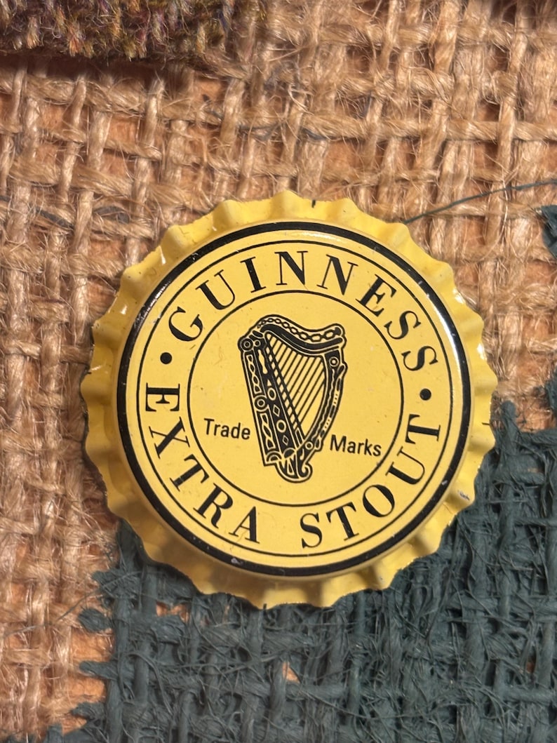 Puede incluir: Un tap&oacute;n de botella de metal amarillo con letras negras que dice "GUINNESS EXTRA STOUT" y un logotipo de arpa negro con las palabras "Trade Marks" debajo.