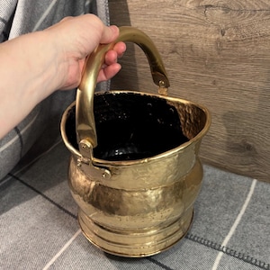 Könnte beinhalten: Ein Vintage-Eimer aus Messing mit gebogenem Griff. Der Eimer hat eine gehämmerten Textur und einen dunklen Innenraum. Der Griff ist mit Scharnieren befestigt. Der Eimer hat eine warme Goldfarbe.