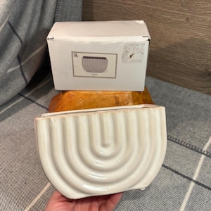 May include: A white ceramic planter with a unique curved design and a ribbed texture. The planter is in the shape of a half-circle.