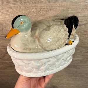 May include: A ceramic duck figurine in a white woven basket. The duck is painted in shades of brown, white, and green with a black head and yellow beak.