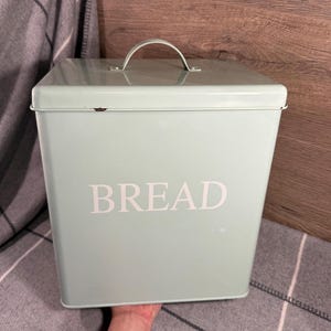 May include: A rectangular, light green bread box with a curved handle on the lid. The word "BREAD" is printed in white on the front of the box. The bread box is made of metal.