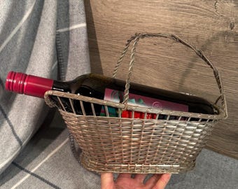 Puede incluir: Una cesta de metal plateado con asa contiene una botella de vino tinto. La botella de vino tiene un tapón burdeos y una etiqueta. La cesta es de metal tejido y está sobre una manta a cuadros gris y blanco.