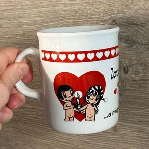 Könnte beinhalten: Weiße Keramik-Tasse mit rotem Herzdesign und einer Reihe roter Herzen am Rand. Die Tasse zeigt ein Cartoon-Paar, das eine Kerze hält. Das Wort "Love" steht auf der Seite.