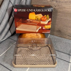 Peut inclure: Un dôme vintage en plastique beige pour aliments et fromages avec un couvercle en verre transparent. Le dôme est posé sur un tissu à motifs marrons. Le dôme est dans sa boîte d'origine, qui est marron avec du texte blanc qui dit "Speise- und Käseglocke Bologna".