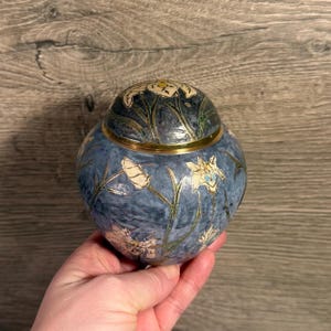 Może przedstawiać: Niebieski dekoracyjny słoik z pokrywką, z kwiatowym wzorem w kolorze białym i złotym. Słoik ma okrągły kształt i złoty pasek pośrodku. Pokrywka jest również ozdobiona kwiatami.