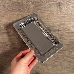 May include: A rectangular, silver-colored metal tray with a polished surface. The tray has decorative openwork along the edges and a beaded border. The tray is held by a hand, and the background is a wooden surface.