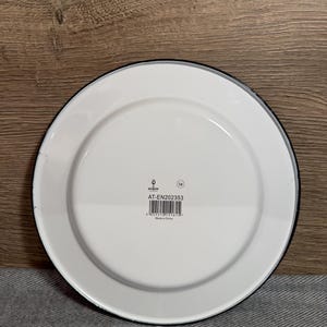 Pode incluir: Um prato redondo de esmalte branco com uma borda preta fina. O centro apresenta uma etiqueta retangular branca com o texto argon, o c&oacute;digo AT-EN202353, um c&oacute;digo de barras e o texto Made in China. O prato tem um acabamento liso e brilhante.