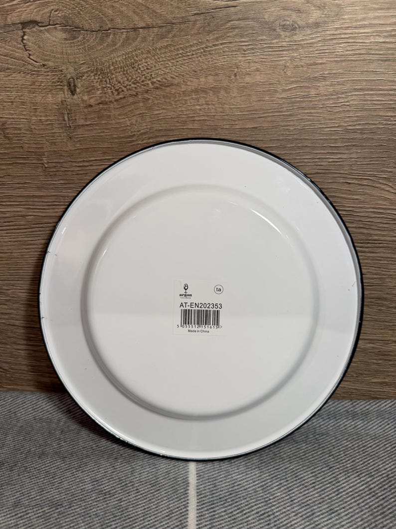 Pode incluir: Um prato redondo de esmalte branco com uma borda preta fina. O centro apresenta uma etiqueta retangular branca com o texto argon, o c&oacute;digo AT-EN202353, um c&oacute;digo de barras e o texto Made in China. O prato tem um acabamento liso e brilhante.