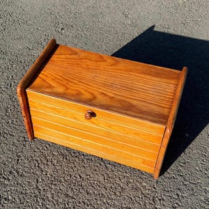Pode incluir: Uma caixa de pão de madeira com um acabamento quente e natural. A caixa apresenta um botão arredondado e um design frontal com ripas. A parte superior e as laterais têm uma aparência lisa e sólida. A caixa de pão tem formato retangular.