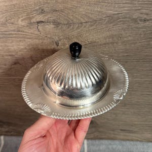 Könnte beinhalten: Silberne Butterdose mit geripptem, gewölbtem Deckel und schwarzem Knauf. Die Schale steht auf einem runden, dekorativen Sockel mit Perlenrand und fächerförmigen Akzenten. Ein klassisches Servierstück.