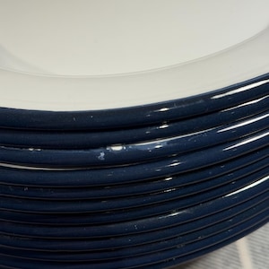 Pode incluir: Uma pilha de pratos redondos de cer&acirc;mica com um centro branco brilhante e uma borda larga azul-marinho escura. As bordas dos pratos apresentam um perfil liso e arredondado, com uma pequena &aacute;rea de desgaste branco na borda azul de um prato.