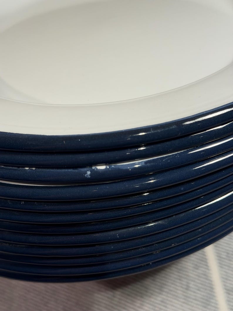 Pode incluir: Uma pilha de pratos redondos de cer&acirc;mica com um centro branco brilhante e uma borda larga azul-marinho escura. As bordas dos pratos apresentam um perfil liso e arredondado, com uma pequena &aacute;rea de desgaste branco na borda azul de um prato.