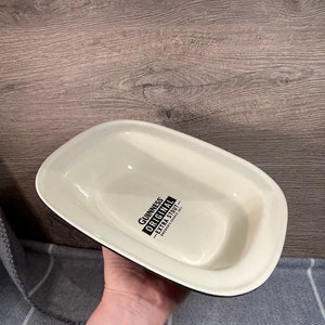 May include: Cream-colored, rectangular serving dish with a black rim. The dish has the words "Guinness Original Extra Stout" printed in black. The dish is held by a person.