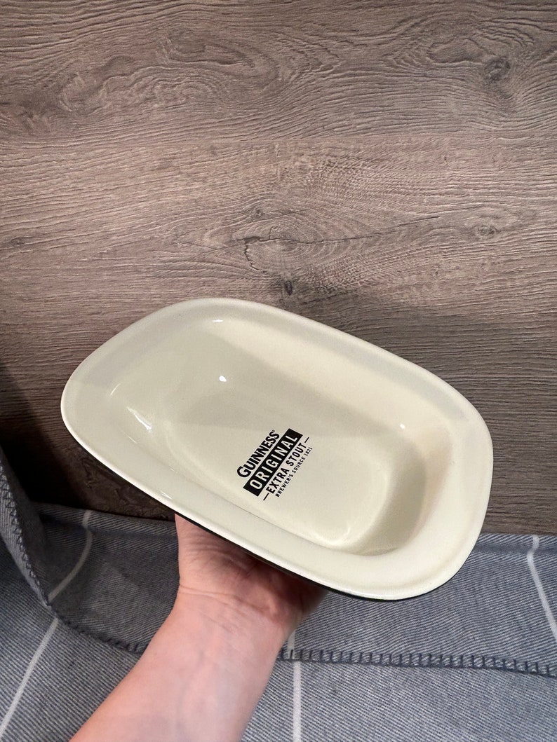May include: Cream-colored, rectangular serving dish with a black rim. The dish has the words "Guinness Original Extra Stout" printed in black. The dish is held by a person.