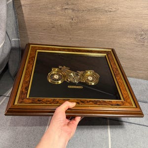 May include: A framed artwork with a gold motorcycle design on a black background. The wooden frame has a dark brown finish and a gold inner border. The motorcycle's wheels and engine are intricately detailed, creating a vintage aesthetic.