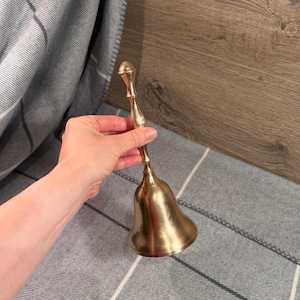 May include: A polished brass hand bell featuring a long, slender, contoured handle and a wide, flared base. The metal surface has a reflective, metallic gold finish with subtle vertical striations visible along the bell's curved body.