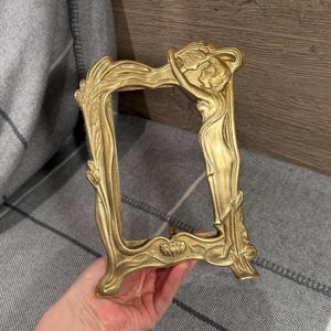 Op de afbeelding: Omlijste goudkleurige fotolijst met Art Nouveau-stijl. De lijst heeft een gebeeldhouwde vrouwelijke figuur en bloemmotieven. De lijst is rechthoekig met een decoratieve basis en bovenkant. De lijst is van metaal.