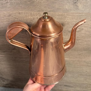 May include: A copper-colored tea kettle with a long spout, a handle, and a lid with a small knob. The kettle has a shiny finish and is likely made of copper. The design is vintage and suitable for brewing tea.