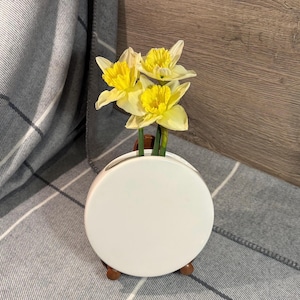 以下が含まれることがあります： 丸い形をした白い陶器の花瓶で、木製のスタンドが付いています。花瓶の上部に開口部が1つあり、3本の黄色の水仙が入っています。