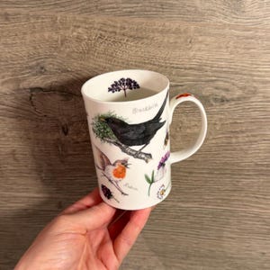 May include: A white ceramic mug featuring detailed illustrations of a blackbird and a robin perched on branches, accompanied by botanical elements and berries. The text Blackbird and Robin is printed in script near the respective birds.