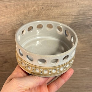 May include: A small, round, ceramic bowl with a light gray interior and exterior. The rim features evenly spaced circular cutouts. A band of tan and white decorative circles encircles the base of the bowl. The bowl is held in a hand.