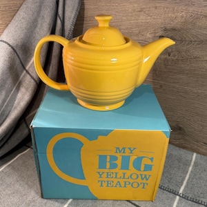 Könnte beinhalten: Eine leuchtend gelbe Teekanne mit Deckel und Henkel steht auf einer blauen Schachtel. Auf der Schachtel steht in Gelb "MY BIG YELLOW TEAPOT". Die Teekanne ist aus Keramik.