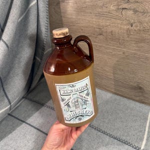 Puede incluir: Una jarra de whisky de cerámica bicolor con la parte superior esmaltada en marrón oscuro y la base en color canela. Incluye un tapón de corcho, un asa lateral y una etiqueta rectangular con el texto WHIMEBRAYER HAND-MADE SOUR MASH WHISKEY.