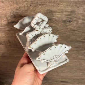 Puede incluir: Una pieza decorativa de cerámica blanca con una base rectangular, tres elementos en forma de abanico y una estructura similar al bambú. Los abanicos y el bambú tienen detalles dorados. Probablemente sea un artículo decorativo para el hogar.