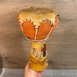 Op de afbeelding: Een kleine handtrommel met een bruinleren trommelvel en een oranje body. De trommel is versierd met zwarte stiksels en witte touwtjes. De basis is geel met een geschilderd ontwerp.