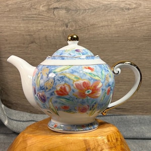 Puede incluir: Una tetera de porcelana blanca con un patrón azul y floral. La tetera tiene un borde dorado y una perilla dorada en la tapa.