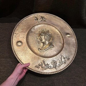 Può includere: Un piatto decorativo rotondo in ottone con un profilo centrale in rilievo di una figura con elmo sopra un cartiglio con il testo SIR GALAHAD. Il piatto presenta una texture martellata, un bordo a corda e decorazioni a volute sul fondo.