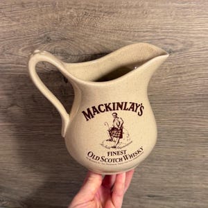 Op de afbeelding: Beige keramische kan met een handvat en schenktuit. Het heeft de tekst "MACKINLAY'S FINEST OLD SCOTCH WHISKY" in bruin, met een illustratie van een man in een kilt. De kan heeft een gespikkelde afwerking.