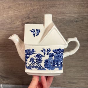 Peut inclure: Une théière en céramique blanche avec un couvercle en forme de maison et une poignée. La théière présente un motif Willow bleu et blanc avec une scène de paysage. Le bec et la poignée sont incurvés. La théière est tenue par une personne.