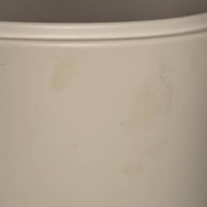 May include: A cylindrical container with a smooth, matte off-white finish. The exterior surface features a subtle, raised horizontal rim near the top edge and shows several faint, irregular light brown stains across the side of the vessel.