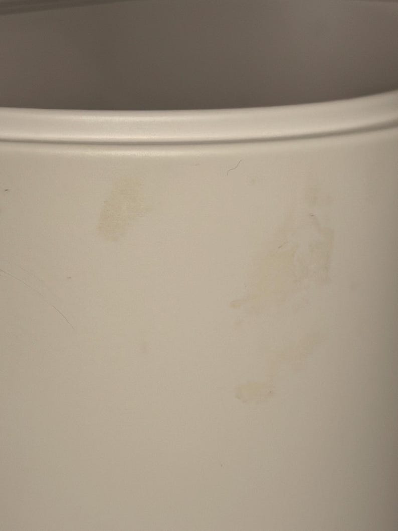 May include: A cylindrical container with a smooth, matte off-white finish. The exterior surface features a subtle, raised horizontal rim near the top edge and shows several faint, irregular light brown stains across the side of the vessel.