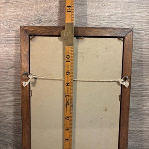 May include: Back of a rectangular wooden picture frame with a beige backing. A wooden ruler is placed vertically against the frame, measuring approximately 16 inches in height. The frame has a string for hanging.