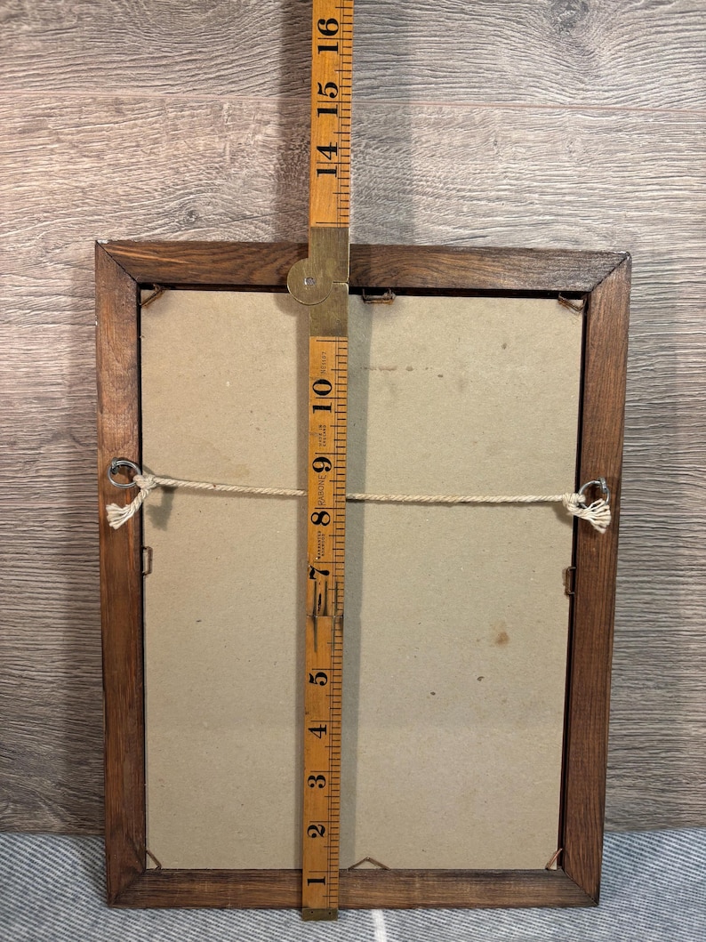 May include: Back of a rectangular wooden picture frame with a beige backing. A wooden ruler is placed vertically against the frame, measuring approximately 16 inches in height. The frame has a string for hanging.