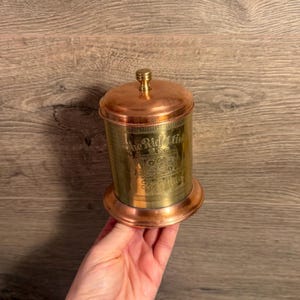 May include: A copper and brass container with a lid and base. The container has the words "The Right Time" etched on the side. The lid has a small knob on top. The container is cylindrical and sits on a round base.