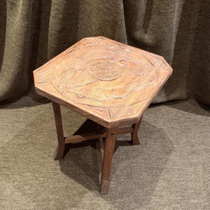 Könnte beinhalten: Ein kleiner, achteckiger Beistelltisch aus Holz mit einer geschnitzten Tischplatte, die ein zentrales kreisförmiges Motiv und geometrische Muster aufweist. Der Tisch steht auf vier geraden Beinen, die durch ein kreuzförmiges Gestell verbunden sind.