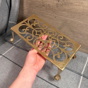May include: A rectangular brass trivet with ornate cut-out designs and four claw feet. The trivet is held in a hand, showcasing its intricate patterns and golden color. The trivet measures approximately 8 inches long and 4 inches wide.