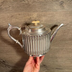 May include: A silver teapot with a fluted body, decorative handle, and spout. The lid has a decorative finial. The teapot is held by a hand against a wooden background. A classic piece for tea service.