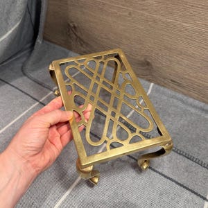 Pode incluir: Um descanso de panela retangular em latão com um padrão intrincado de treliça vazada de laços e linhas entrelaçadas. O suporte de metal é apoiado por quatro pernas curvas, proporcionando uma plataforma elevada com acabamento dourado.